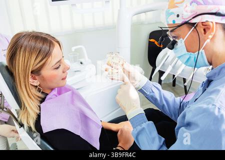 Dentiste portant un masque chirurgical et une loupe montrant un modèle de prothèse dentaire à une jeune patiente assise sur le fauteuil du dentiste dans une clinique dentaire lumineuse Banque D'Images