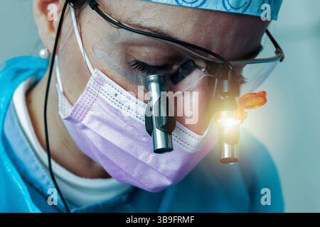 Dentiste femme portant une casquette chirurgicale, des loupes avec lumière et un masque rose, effectuant un examen dentaire approfondi dans un cadre de clinique dentaire moderne Banque D'Images