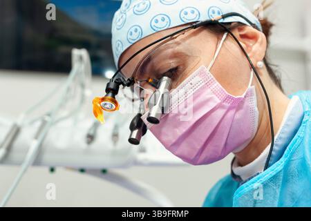 Dentiste femme portant une casquette chirurgicale, des loupes avec lumière et un masque rose, effectuant un examen dentaire approfondi dans un cadre de clinique dentaire moderne Banque D'Images