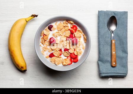 Bol gris rempli de céréales et de lait, surmonté de fraises et de framboises, accompagné d’une banane et d’une cuillère sur une serviette bleue sur une table en bois blanc Banque D'Images