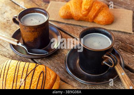 Deux tasses de café noir fumantes sont assises sur une table en bois rustique à côté d'un croissant et d'un saccottino, créant une scène de petit déjeuner confortable et accueillante Banque D'Images