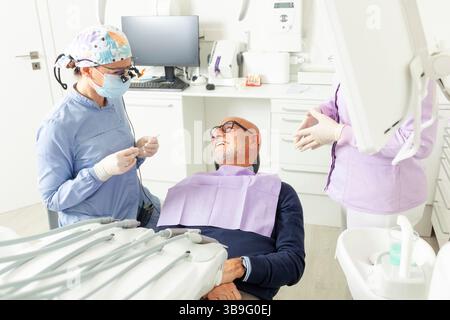 Dentiste portant des loupes préparant des outils pour examiner un homme âgé assis dans un fauteuil dentaire, pendant qu'une infirmière l'assiste Banque D'Images