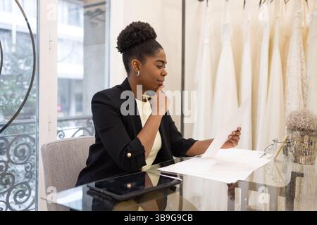 Femme d'affaires professionnelle dans un magasin de mariée examinant les documents attentivement. Vêtue d'une tenue habillée, elle est assise à un bureau en verre, entourée d'élégants Banque D'Images