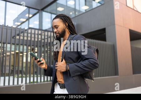 Homme d'affaires portant un blazer et un sac à dos, marchant à l'extérieur d'un immeuble de bureaux moderne tout en vérifiant son smartphone. Parfait pour les thèmes d'affaires, Mo Banque D'Images