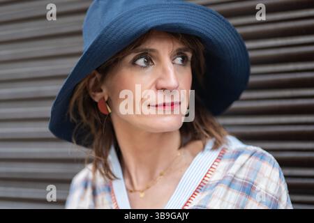Une femme de mode portant un chapeau bleu et une chemise à carreaux se tient sur un fond sombre, exsudant une aura réfléchie et élégante. Ses boucles d'oreilles rouges et makeu Banque D'Images