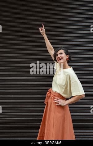Une femme de mode dans une chemise beige et une jupe orange sourit et pointe vers le haut sur un fond texturé, véhiculant positivité et aspiration dans une CA Banque D'Images