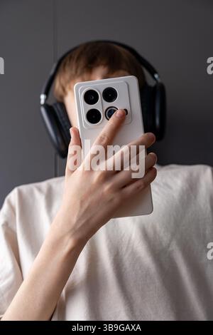 Un adolescent portant de grands écouteurs noirs tient un smartphone avec un étui blanc devant son visage. Le gros plan capture un moment moderne du numérique Banque D'Images