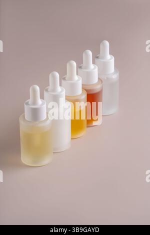 Ensemble de cinq flacons compte-gouttes translucides avec différents contenus de couleur sur un fond beige doux, reflétant la lumière et de légères variations. Banque D'Images