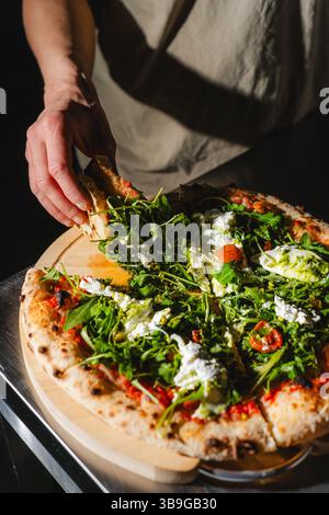 Une personne attrape une tranche de pizza garnie de roquette fraîche et de fromage crémeux. La pizza est placée sur une planche de bois avec un éclairage chaud améliorant son de Banque D'Images