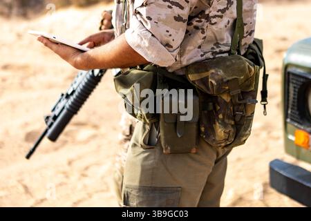 Un soldat vêtu d'un équipement militaire tactique vérifie un appareil numérique tout en tenant un fusil. La scène capture un moment de stratégie et de concentration dans un désert Banque D'Images