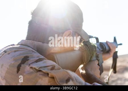 Un soldat en camouflage vise un fusil dans un paysage désertique ensoleillé. Le soleil éclatant crée un éclat de lentille saisissant, mettant en évidence le contexte militaire et Banque D'Images