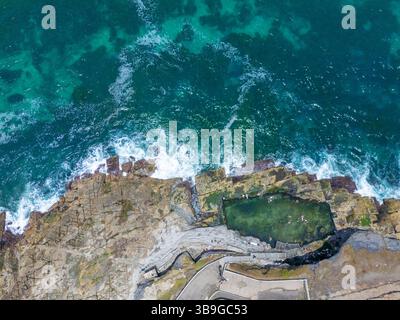 Vue aérienne de la côte de la Nouvelle-Galles du Sud mettant en valeur un bassin rocheux naturel entouré de rochers escarpés. Les vagues bleues vibrantes de l'océan s'écrasent contre le sho Banque D'Images