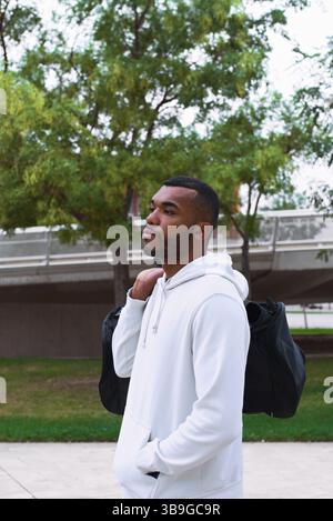 Portrait d'un jeune athlète masculin noir dans un sweat à capuche blanc portant un sac de sport à l'extérieur. Le cadre comprend la verdure et l'architecture moderne dans le Banque D'Images