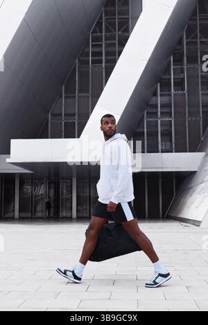 Un athlète noir confiant est capturé en mouvement, portant un sweat à capuche et un short dans un cadre urbain. L'architecture moderne complète sa dynamique Banque D'Images
