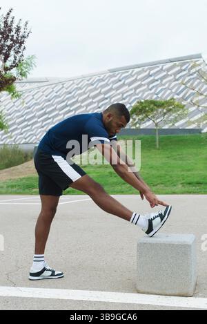 Athlète masculin noir en vêtements de sport s'étire à l'extérieur, se préparant à une séance d'entraînement près d'un bâtiment moderne. Mettre l'accent sur la forme physique, le dévouement et l'athlétisme. Banque D'Images