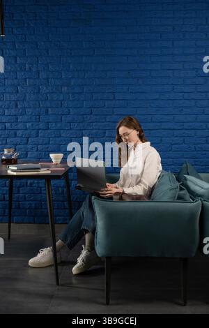 Une femme en tenue décontractée travaille sur un ordinateur portable, assise sur un canapé sarcelle contre un mur de briques bleues. Une table avec thé et cahiers complète le confortable atmos Banque D'Images