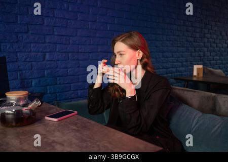 Une femme dans une tenue noire savoure un thé chaud dans un bar élégant avec un mur de briques bleues. Une théière et un téléphone sont sur la table, créant une atmosphère détendue Banque D'Images