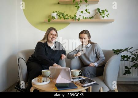 Deux femmes sont assises dans un café confortable, appréciant le café et la conversation. Les ordinateurs portables et les cahiers sont sur la table, suggérant un mélange de travail et de loisirs dans un rela Banque D'Images