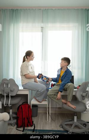 Un garçon et une fille partagent un moment avec une main robotisée. Le cadre est une salle confortable et lumineuse remplie de matériel éducatif qui symbolisent la créativité et Banque D'Images