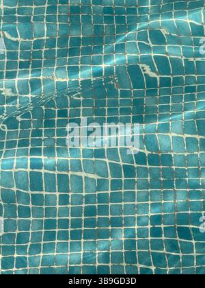 Une vue sereine sur la surface carrelée bleue d'une piscine, avec la lumière du soleil créant des ondulations et des reflets doux. Le mouvement de l'eau ajoute un texte apaisant Banque D'Images