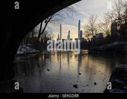 Un coucher de soleil serein illumine les gratte-ciel de New York, vu de l'autre côté d'un étang gelé sous un pont à Central Park. Le paysage de fin d'hiver offre un y calme Banque D'Images