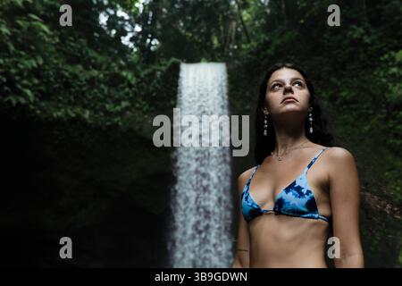 Une femme en bikini bleu se tient devant une cascade majestueuse entourée d'une végétation luxuriante, capturant un moment de tranquillité et de beauté naturelle Banque D'Images