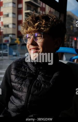 Un jeune homme aux cheveux bouclés et aux lunettes sourit assis dans un bus de la ville. La lumière du soleil met en valeur leur visage, ajoutant de la chaleur et un sentiment de contentement. Banque D'Images