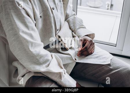 Homme à la mode portant une chemise en lin et un sac bandoulière, assis près de la fenêtre, exsudant une ambiance minimaliste et élégante. Mettant l'accent sur le confort et l'élégance dans un cadre intérieur serein Banque D'Images