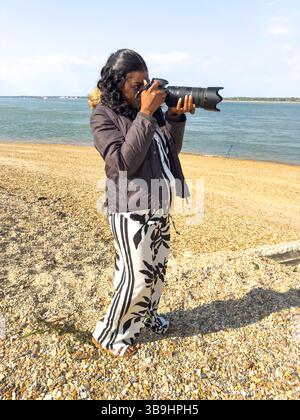 Calshot Hampshire Angleterre Royaume-Uni. 27.04.2025. Femme africaine utilisant un appareil photo numérique reflex numérique lors d'une visite à la plage de Calshot dans le Hampshire Royaume-Uni Banque D'Images