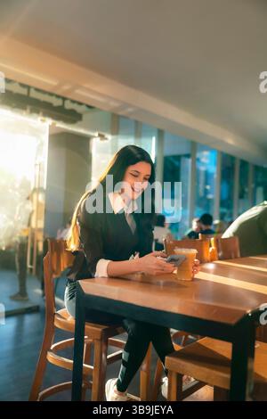 Jeune femme latina souriant tout en vérifiant son téléphone et tenant du café glacé dans un café à Mexico pendant l'heure dorée. Banque D'Images