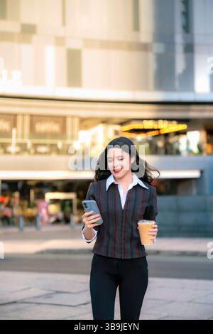 Souriante jeune femme latina dans une tenue décontractée d'affaires marchant à l'extérieur à Mexico en tenant le café et en vérifiant son téléphone. Banque D'Images