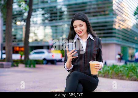 Femme Latina dans une tenue décontractée formelle est assis à l'extérieur sur Reforma, souriant tout en vérifiant son téléphone et tenant un café glacé à Mexico. Banque D'Images