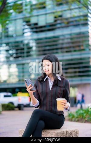 Femme Latina dans une tenue décontractée formelle est assis à l'extérieur sur Reforma, souriant tout en vérifiant son téléphone et tenant un café glacé à Mexico. Banque D'Images