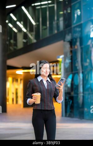 Femme Latina dans une tenue décontractée formelle est assis à l'extérieur sur Reforma, souriant tout en vérifiant son téléphone et tenant un café glacé à Mexico. Banque D'Images