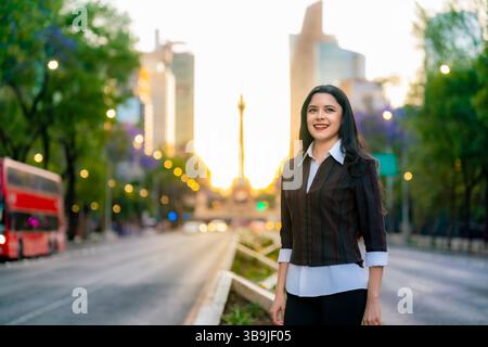 Jeune femme latina avec les cheveux longs et la tenue décontractée d'affaires se tient en toute confiance sur l'avenue Reforma pendant le coucher du soleil à Mexico. Banque D'Images