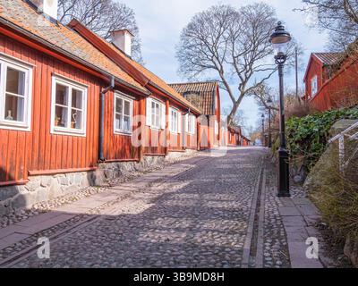 Une rue pavée pittoresque bordée de charmantes maisons en bois rouge et d'arbres, créant une atmosphère sereine. La scène capture un d paisible et ensoleillé Banque D'Images