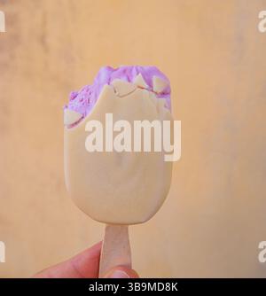 Une main tenant une barre de crème glacée crémeuse avec un dessus violet et enrobage de chocolat blanc sur un fond jaune texturé. Banque D'Images