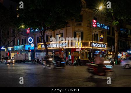 Une scène nocturne animée au Vietnam montre un McDonald's illuminé et la circulation animée. Le paysage urbain est vivant avec l'énergie de la ville. Banque D'Images