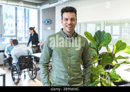 Portrait d'un homme d'affaires souriant debout dans un bureau moderne avec ses collègues handicapés travaillant en arrière-plan Banque D'Images