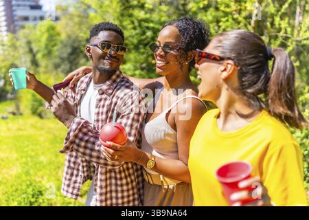 Trois amis marchent dans un parc, appréciant des boissons et la compagnie de l'autre par une journée ensoleillée Banque D'Images