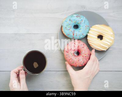 Mains de femme avec café et beignets assortis sur une table en bois gris. Vue de dessus de beignets colorés et de café sur fond en bois gris Banque D'Images