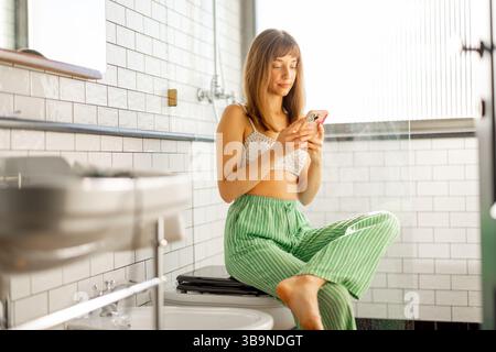 Une jeune femme est assise confortablement dans une salle de bain moderne, absorbée dans son téléphone pendant un matin calme une tranche de routine numérique à la lumière naturelle Banque D'Images