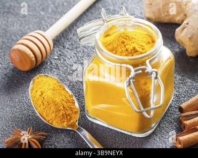 Poudre de curcuma séchée dans un pot en verre et une cuillère sur fond de ciment noir. Ingrédients pour lait doré, latte curcuma ou boisson détox - miel balancier, gi Banque D'Images