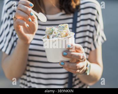 Crème glacée roulée dans la tasse de cône dans les mains de la femme. Femme en robe rayée tient une tasse de cône avec la crème glacée roulée de banane kiwi de style thaïlandais Banque D'Images