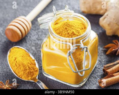 Poudre de curcuma séchée dans un pot en verre et une cuillère sur fond de ciment noir. Ingrédients pour lait doré, latte curcuma ou boisson détox - miel balancier, gi Banque D'Images