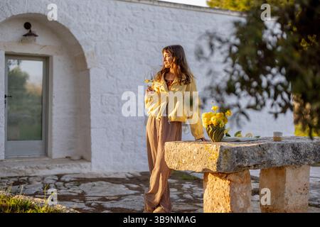 Une femme dans une veste jaune profite d'un verre de vin à l'extérieur d'une maison trullo blanche au coucher du soleil, baignée de lumière dorée, entourée de citrons et de fleurs printanières sur une table en pierre rustique Banque D'Images