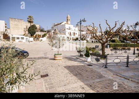 Place de la ville ensoleillée à Alberobello, Italie, avec des rues pavées, des oliviers en pots, des platanes taillés et des toits de trulli emblématiques en arrière-plan. Une scène matinale tranquille Banque D'Images