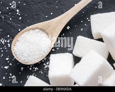 Fond de cubes de sucre et de sucre dans la cuillère. Sucre blanc sur fond noir. Cubes de sucre et granulés dans une cuillère en bois Banque D'Images