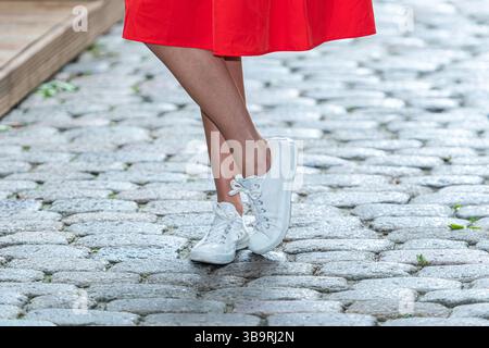 Une femme se tient debout sur une rue pavée portant une robe rouge fluide et des baskets blanches. Banque D'Images