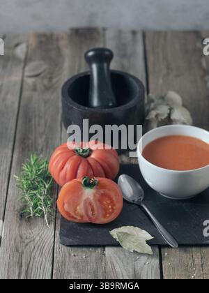 Photo horizontale d'un bol blanc de gazpacho sur une ardoise noire accompagnée de divers ingrédients et ustensiles de cuisine sur une table en bois. Banque D'Images